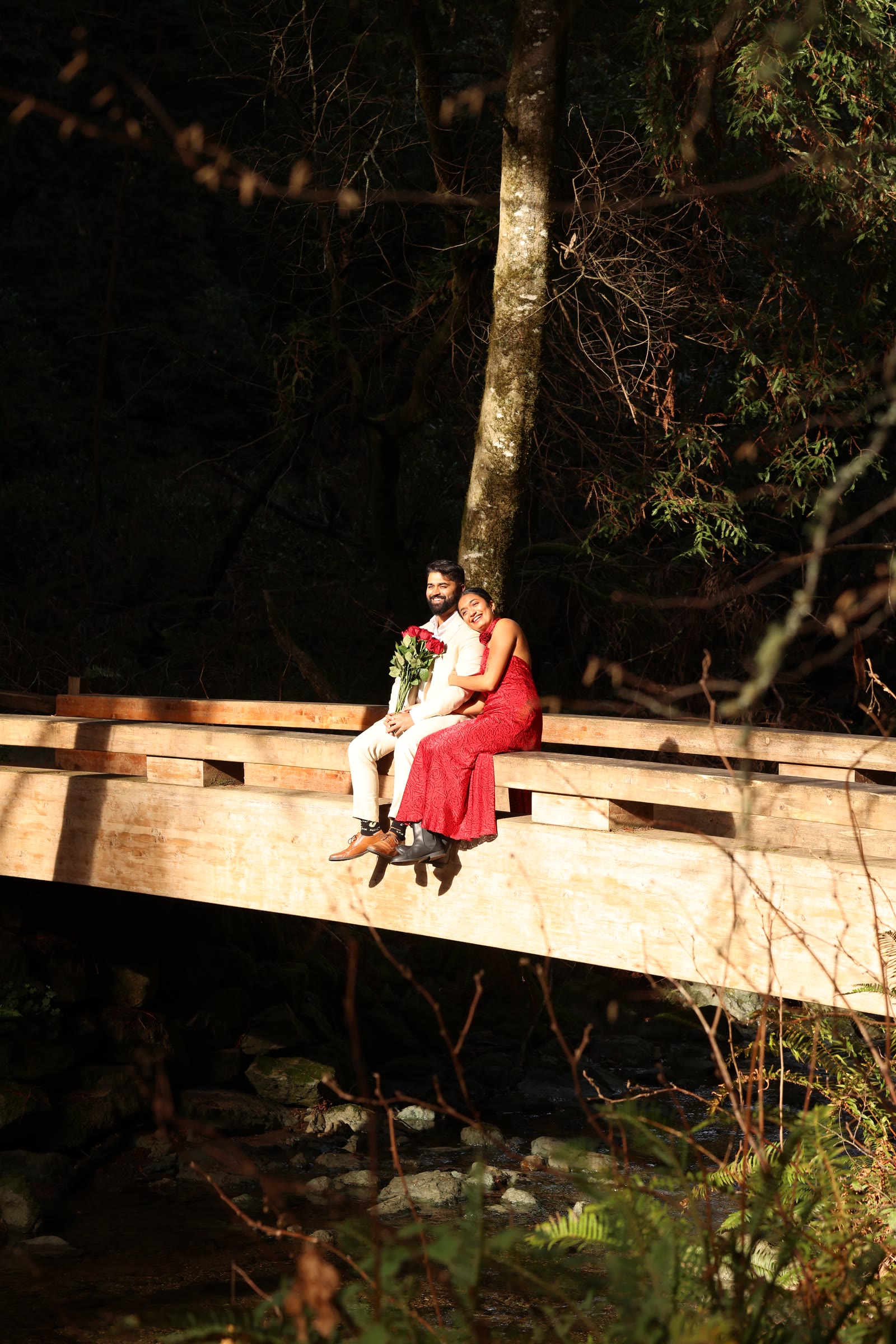 Nish and Pooja sitting on a wooden bridge in a forest, Pooja holding red roses